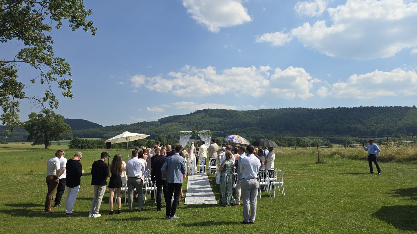 Ceremonia na łące od tyłu: goście stoją, łuk, wzgórza, błękitne niebo